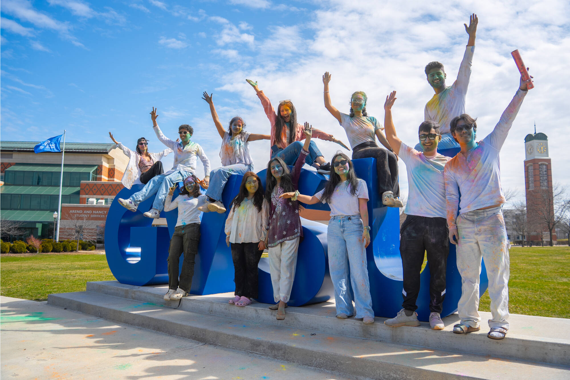 A group of students in white clothes that have been covered in colored powder sitting on the GVSU letters on the Valley campus.